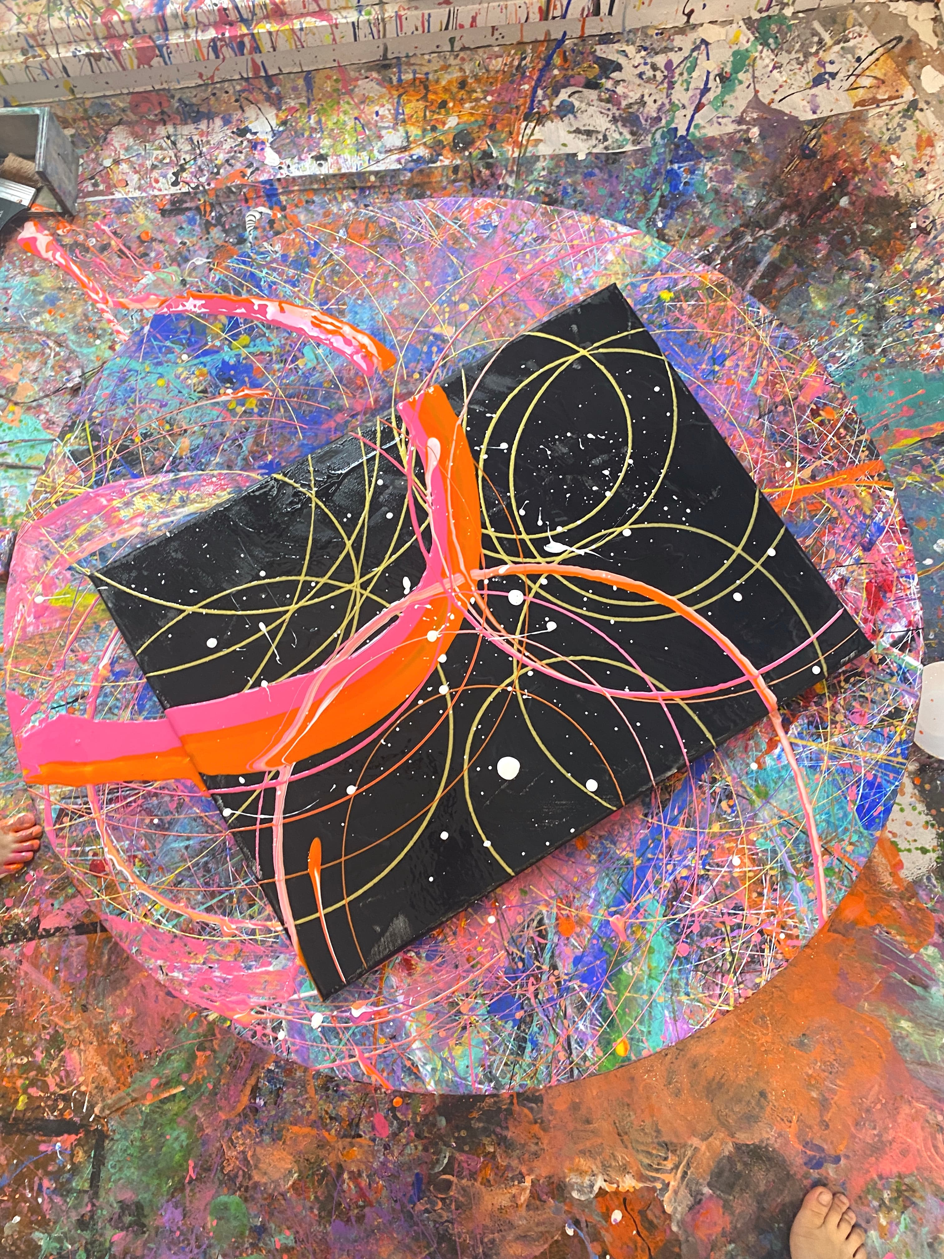 A black canvas on a paint-covered spin table, marked with yellow arcs, white dots and thick pink and orange streaks.