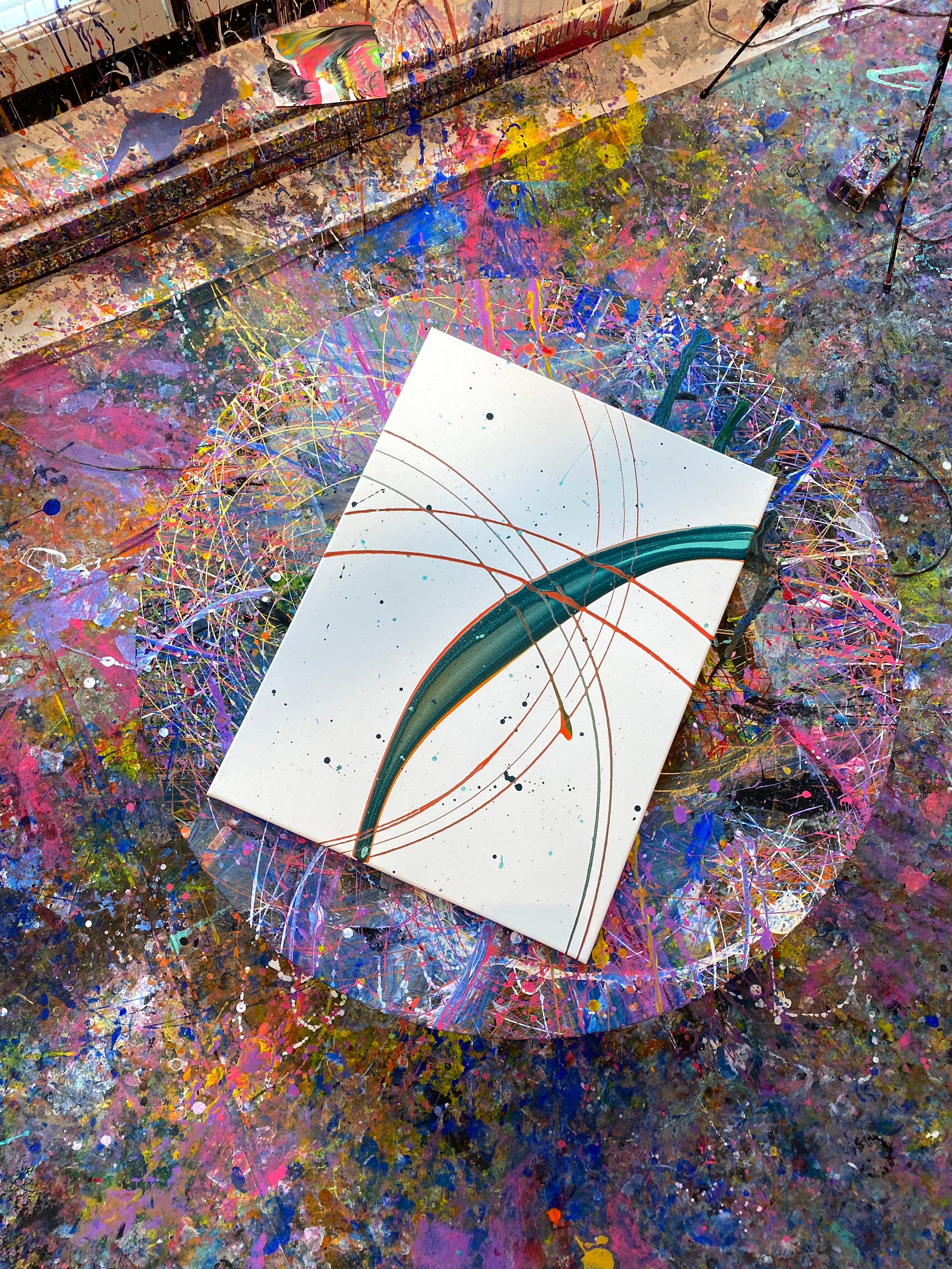 A white canvas on a spin table, marked with thin orange arcs, black speckles and one thick teal stroke.