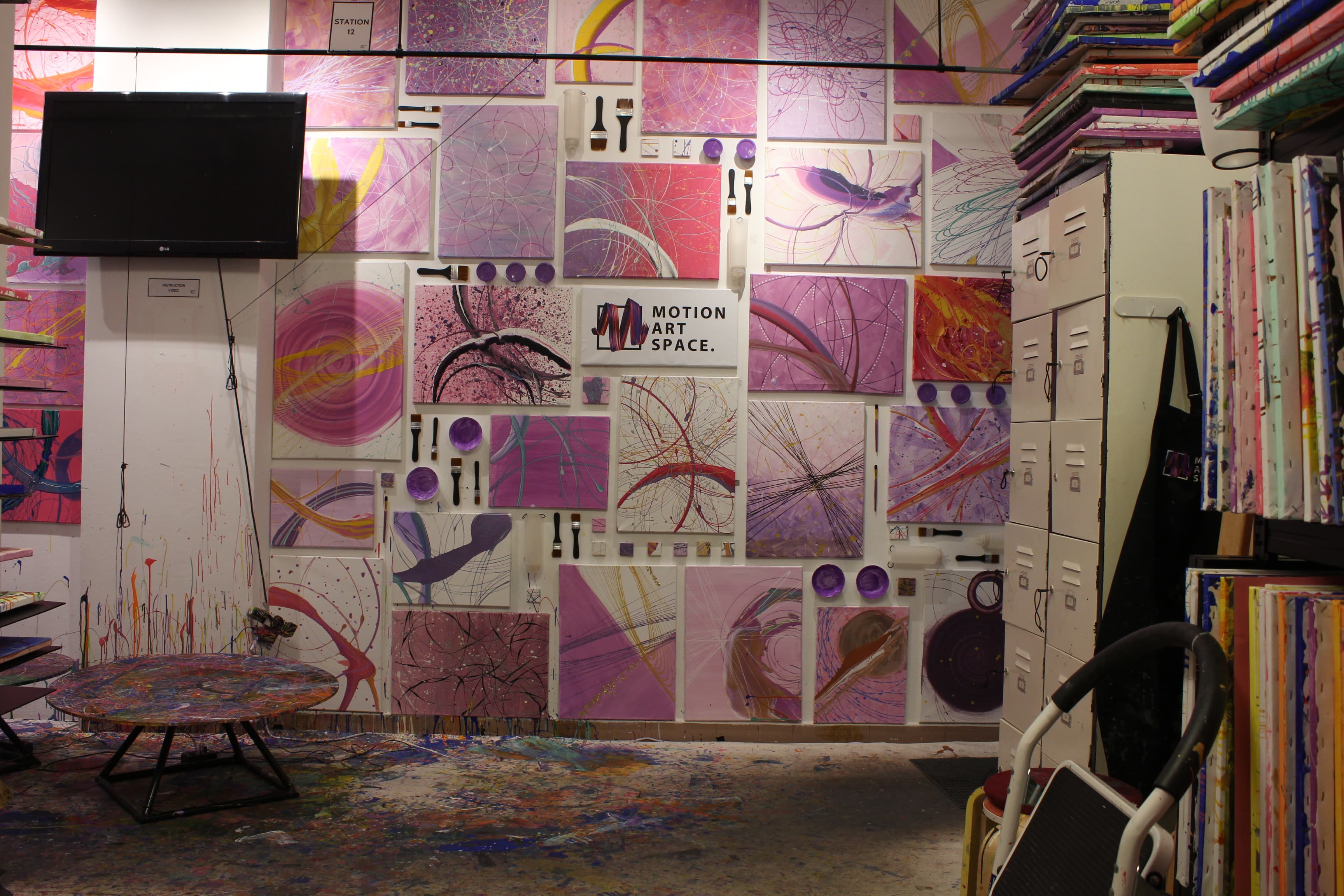 A studio room of pink and purple canvases on the wall above a paint-splattered floor and round table.
