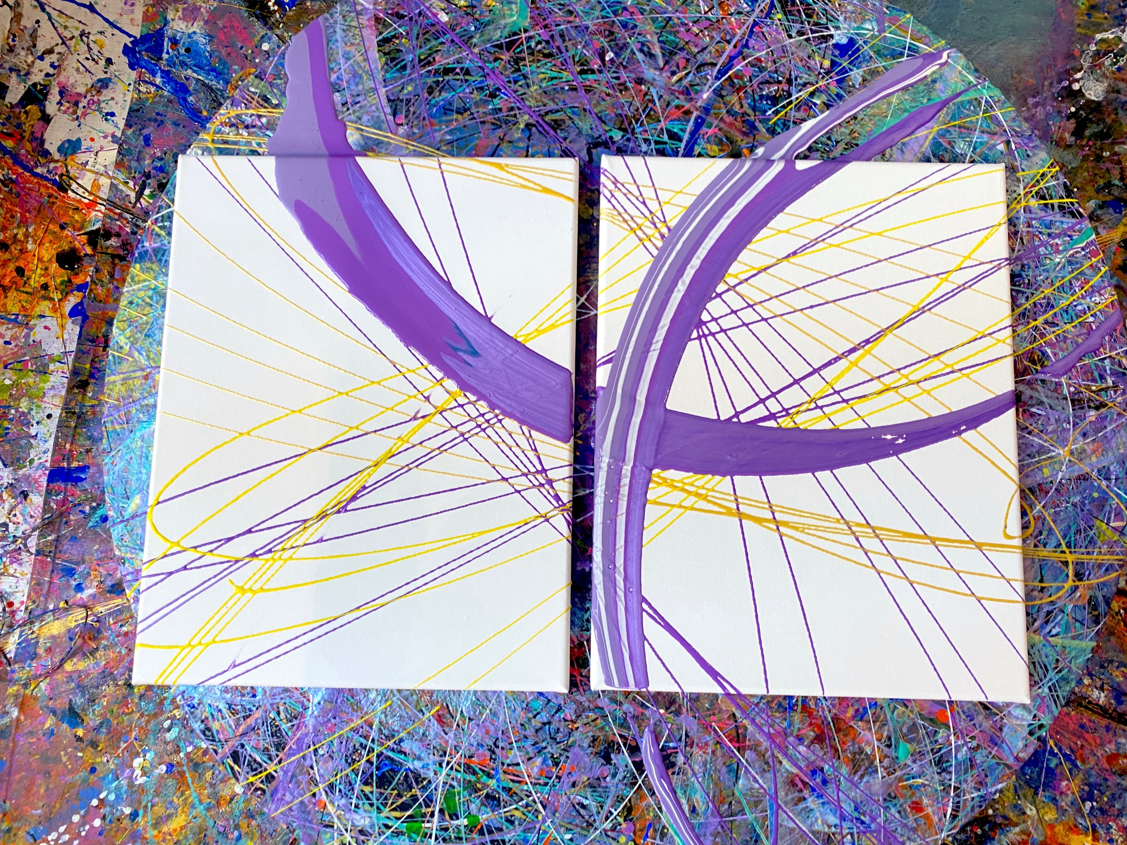 Two white square canvases side by side on a paint-splattered floor, each with purple curved strokes and thin yellow lines.